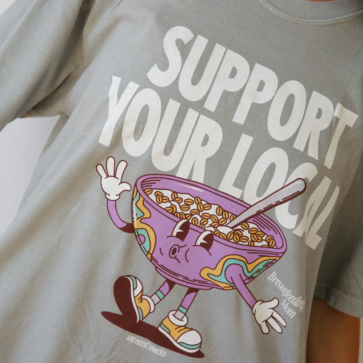 [main] A model wearing a light sage t-shirt featuring the slogan Support Your Local Breastfeeding Mom and a bright cereal bowl graphic. 