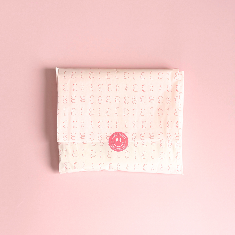 [main] Light Pink tissue paper featuring hot pink shapes of different boobs across the tissue paper to represent breastfeeding, featuring our signature Use Your Voice Smiley Face sticker in the center. 
